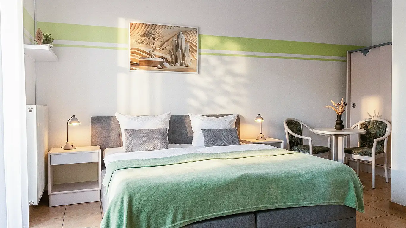 Helles Schlafzimmer mit Doppelbett, grünem Bettüberwurf, zwei Nachttischen mit Lampen und kleinem Tisch mit zwei Stühlen.