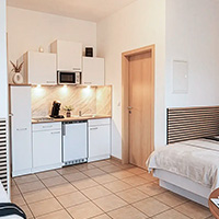 Kleiner Küchenbereich mit weißen Schränken, Mikrowelle, Kaffeemaschine und Geschirrspüler neben einem Bett mit weißer Bettdecke und Holztür.