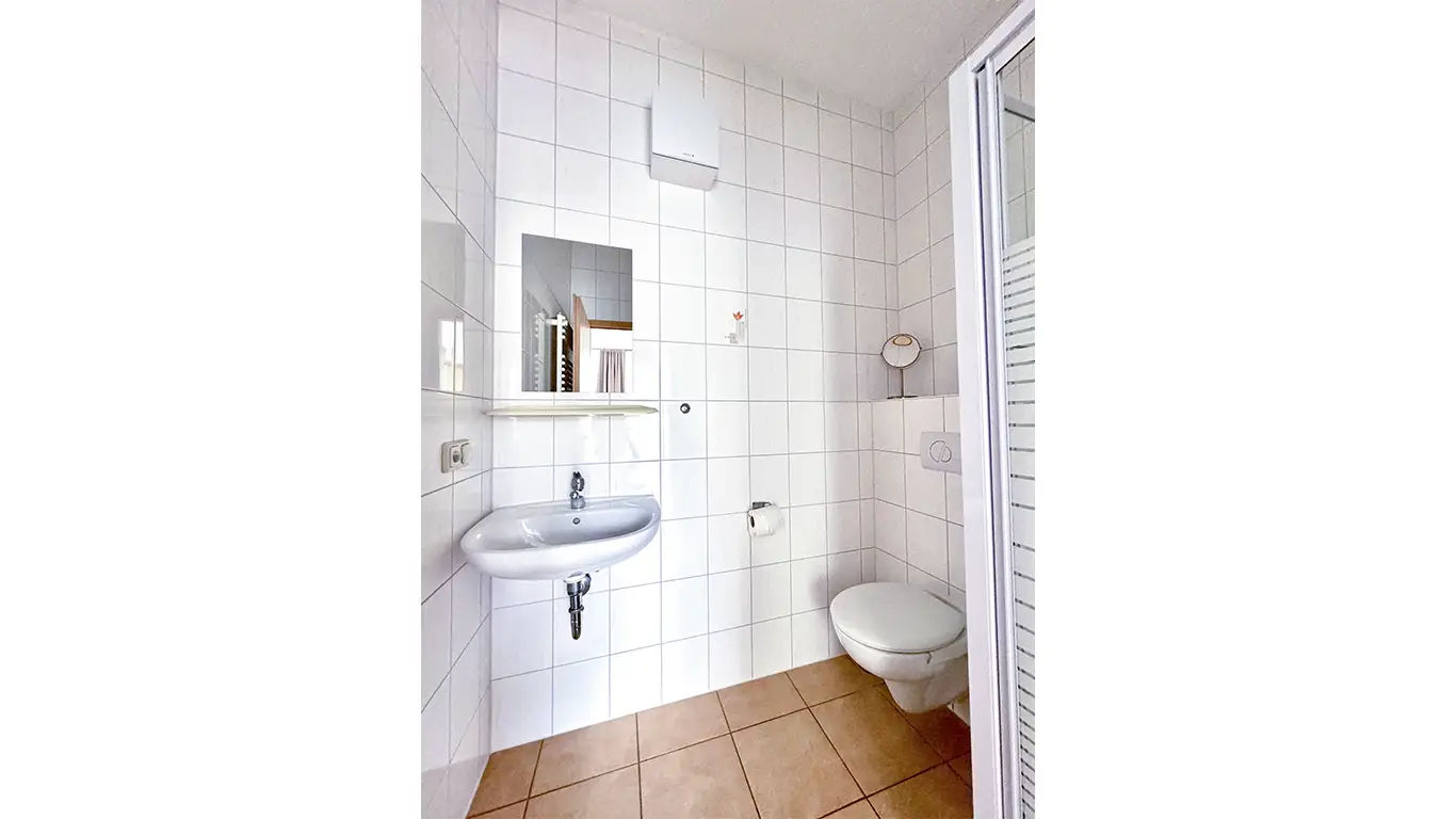 Kleines Badezimmer mit Wand-WC, weißem Waschbecken, Spiegel und hellbraunen Bodenfliesen.