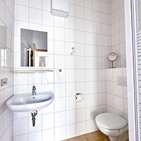 Kleines weiß gekacheltes Badezimmer mit Waschbecken, Toilette, Spiegel und Wandregal.