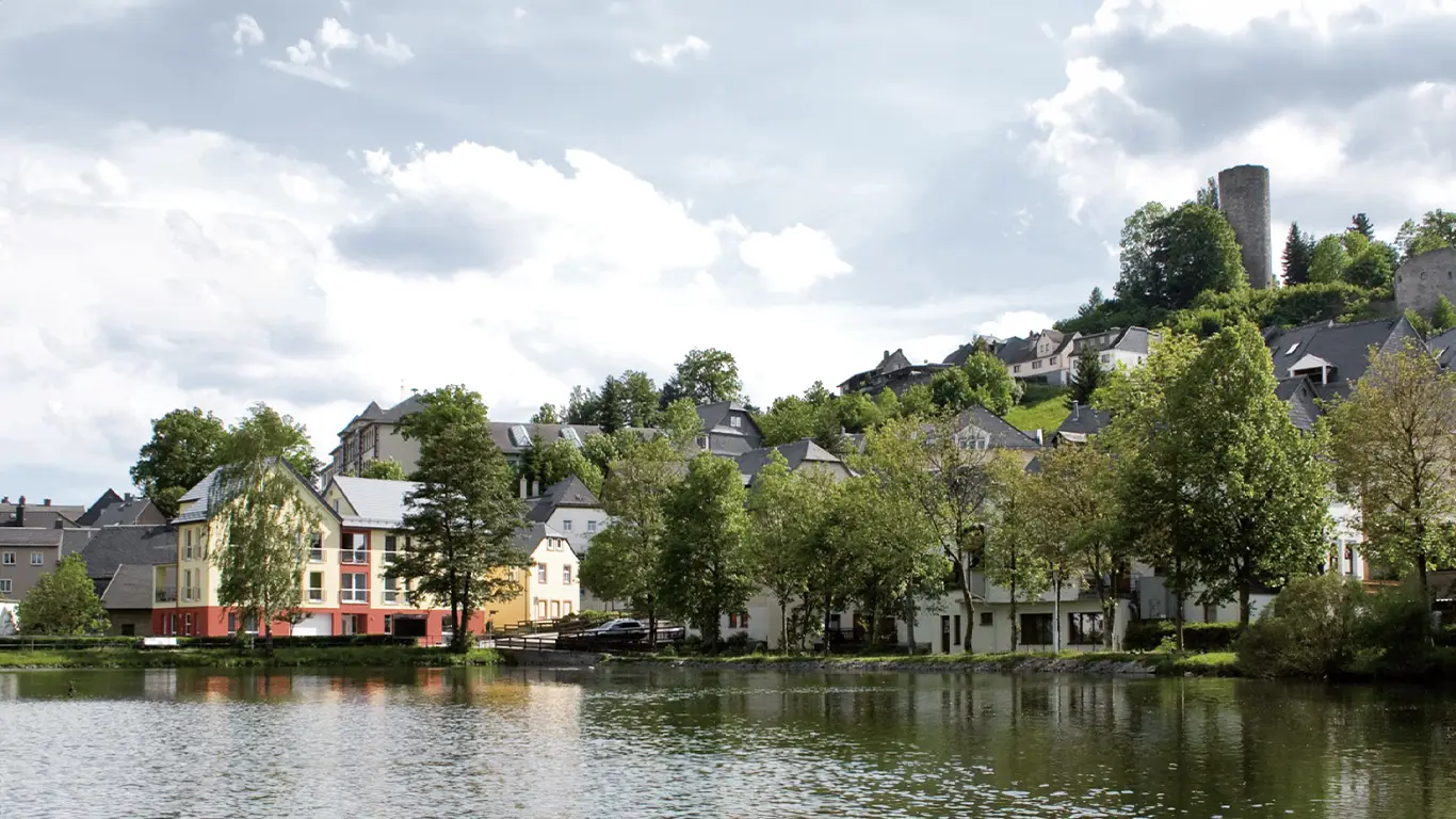 Blick auf einen ruhigen Teich mit Bäumen und bunten Häusern am Ufer, im Hintergrund ein Hügel mit historischen Burgruinen.