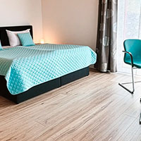 Schlafzimmer mit Doppelbett, türkisfarbenem Steppbett und passendem Stuhl neben einem Fenster mit Vorhängen.