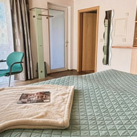 Schlafzimmer mit grünem gestepptem Bett, darauf eine gefaltete Decke und eine Zeitung, im Hintergrund Türen, ein grüner Stuhl und ein Spiegel.