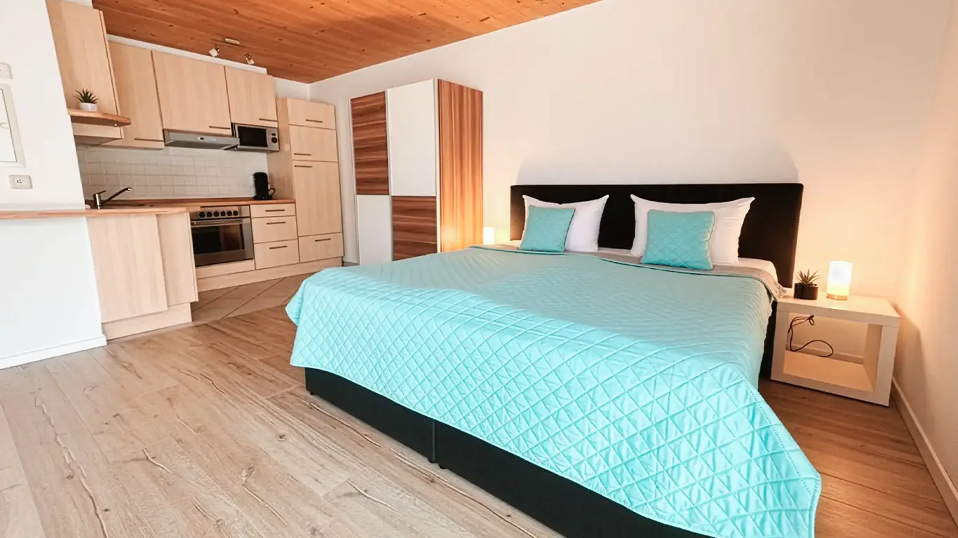 Modern eingerichtetes Zimmer mit Doppelbett, türkisfarbener Bettdecke, Küchenzeile und holzverkleidetem Decken.