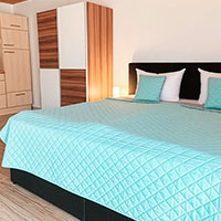 Schlafzimmer mit Doppelbett, hellblauer Steppdecke, zwei türkisfarbenen Kissen und Kleiderschrank aus Holz.