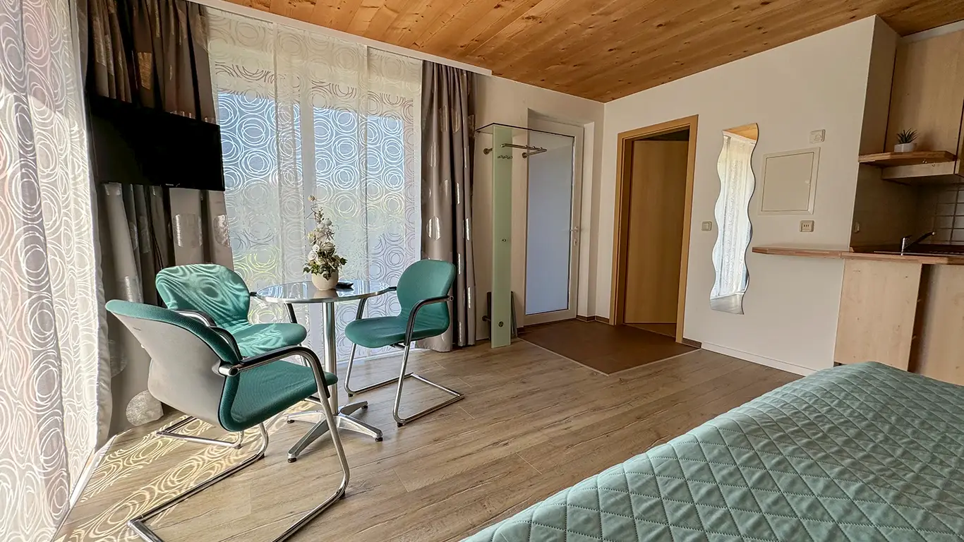 Helles Zimmer mit Holzbalkendecke, zwei grünen Stühlen um einen kleinen runden Tisch, großem Fenster mit transparenten Vorhängen, Wandfernseher und Bett mit grünem Steppüberwurf.