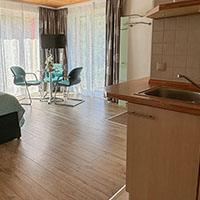 Heller Raum mit Küchenzeile, kleinem runden Esstisch mit vier Stühlen und großen Fenstern mit halbtransparenten Vorhängen.