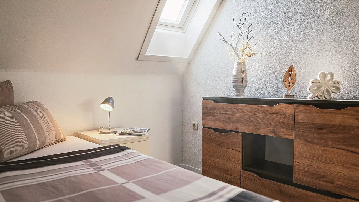 Helles Schlafzimmer mit einem Bett neben einem Nachttisch mit Lampe und einem niedrigen Holzschrank unter einem Dachfenster.