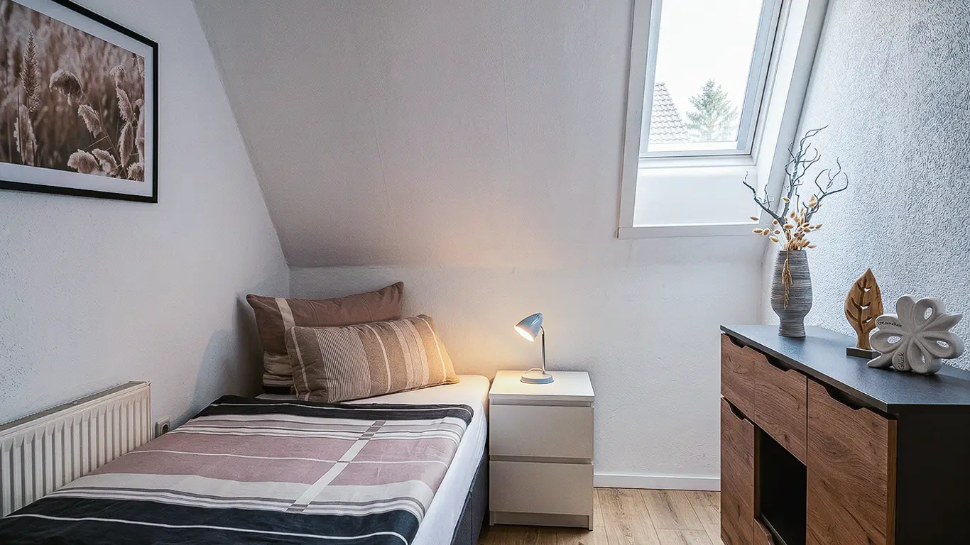 Kleines Schlafzimmer mit Einzelbett, Nachttisch mit Lampe, Dachfenster und Kommode mit Vase und Dekoration.