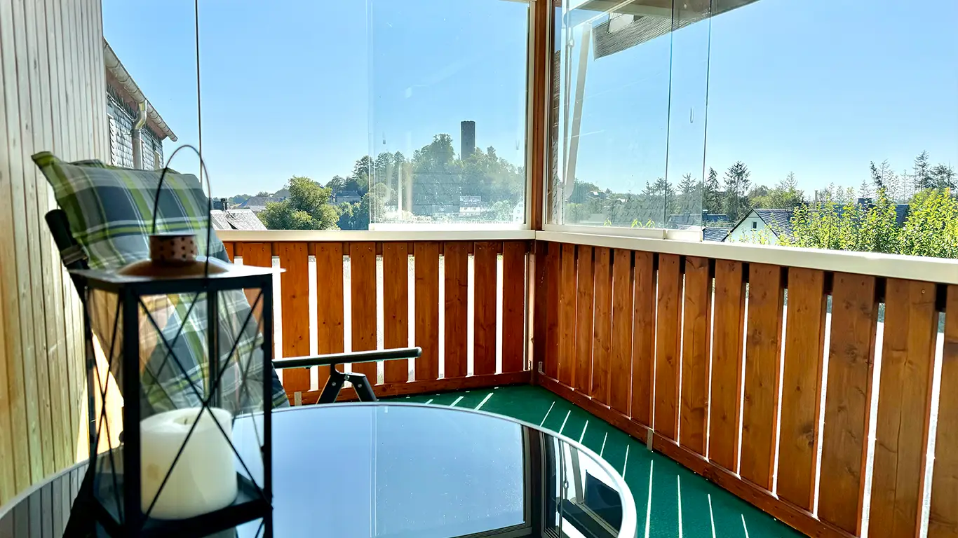 Balkon mit Holzgeländer, grün kariertem Stuhl, Laterne mit Kerze und Blick auf einen Turm bei klarem Himmel.