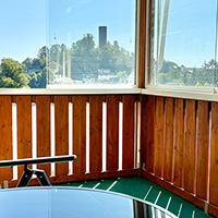 Blick von einem Balkon mit Holzgeländer auf grüne Bäume und blauen Himmel.