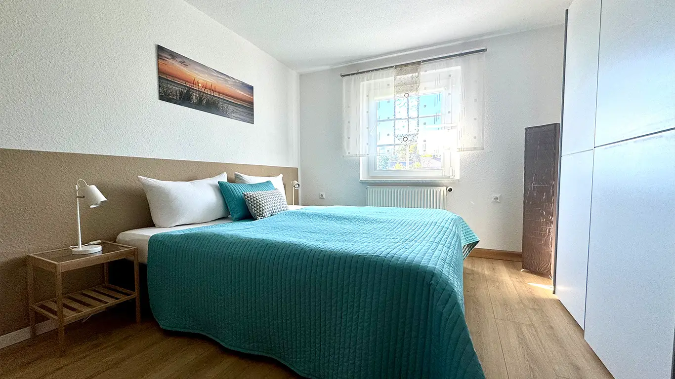 Helles Schlafzimmer mit großem Bett, türkisfarbenem Bettüberwurf, zwei weißen Kissen, Nachttisch mit Lampe und Fenster mit heller Gardine.