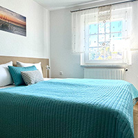 Hell eingerichtetes Schlafzimmer mit großem Fenster, blauem Bettüberwurf und Landschaftsbild an der Wand.