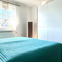 Hell eingerichtetes Schlafzimmer mit grünem Bettbezug, weißem Kleiderschrank und Fenster mit weißen Vorhängen.