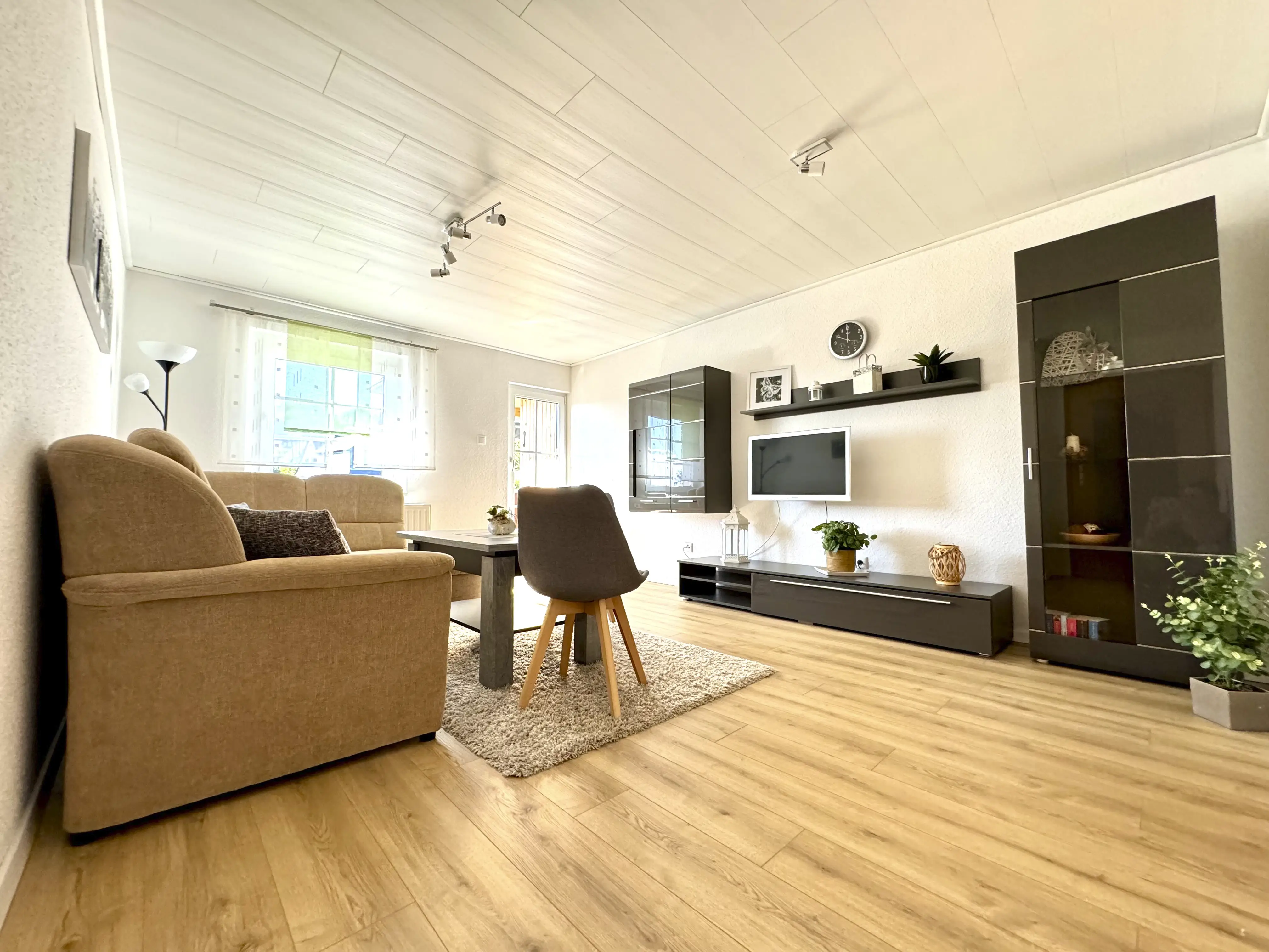 Helles Wohnzimmer mit beige Sofa, grauem Stuhl, Holzboden und moderner schwarzer Wandmöbelwand mit Fernseher und Pflanzen.