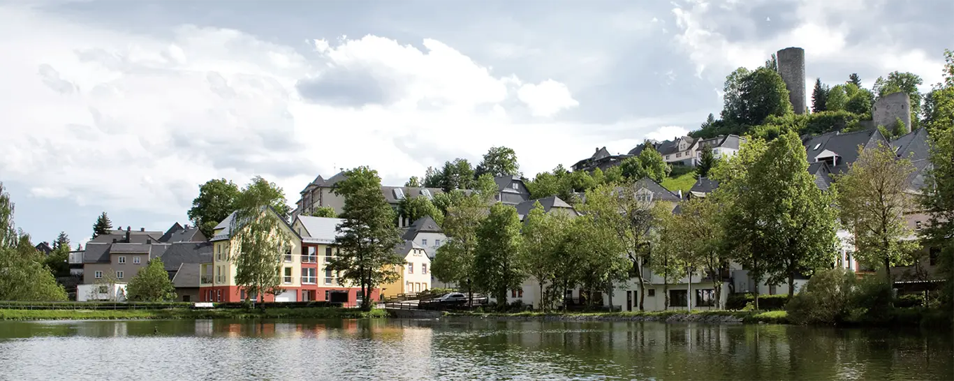 Dorf am Ufer eines ruhigen Sees mit Bäumen und Burgruine auf einem Hügel unter bewölktem Himmel.