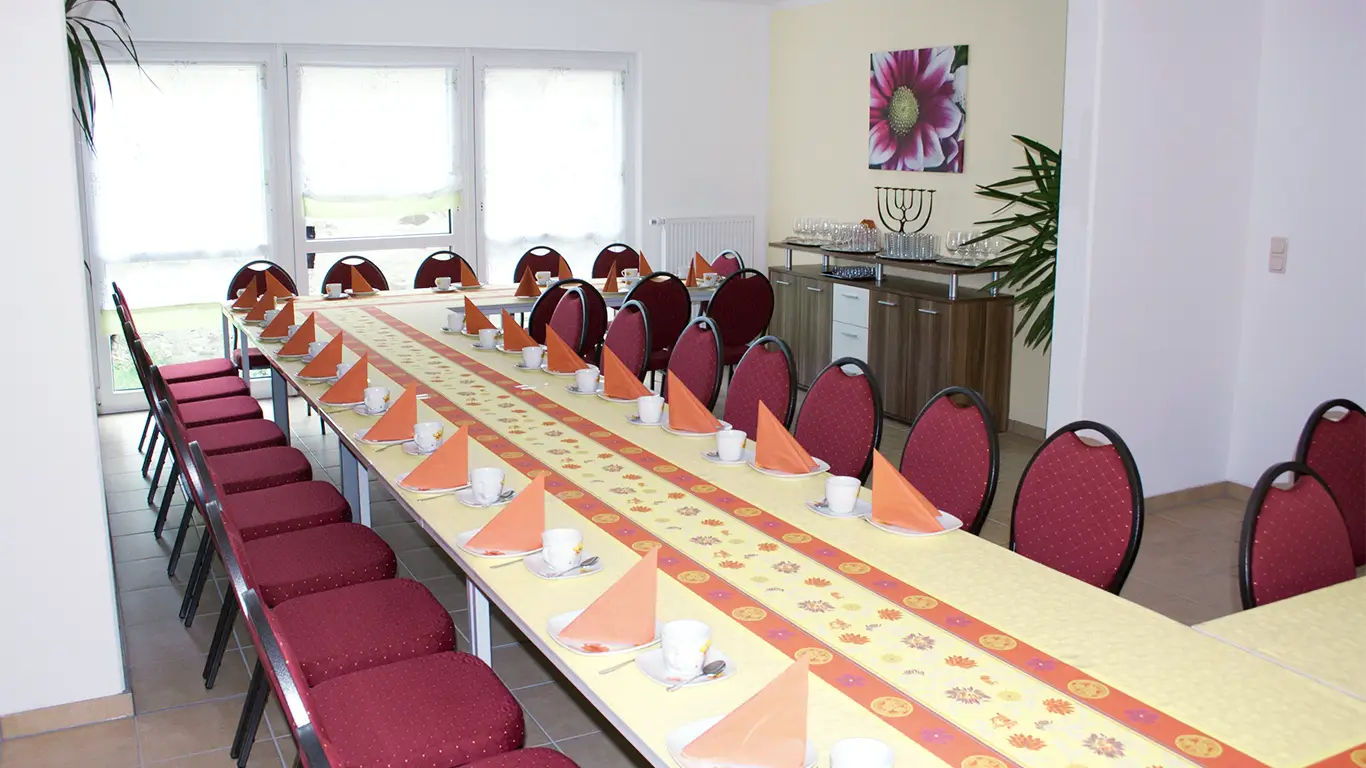 Langer gedeckter Konferenztisch mit roten Stühlen, orangefarbenen Servietten und weißen Tassen in einem hellen Raum.